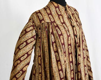 May include: A long-sleeved dress with a paisley pattern in burgundy and tan. The dress has a high collar and button closures. The fabric features vertical stripes of paisley designs. The dress is displayed on a mannequin.