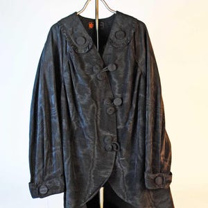May include: A black vintage jacket with a unique design. The jacket features a ruffled collar, decorative buttons, and long sleeves with buttoned cuffs. The fabric has a subtle sheen, and the jacket is displayed on a metal hanger.
