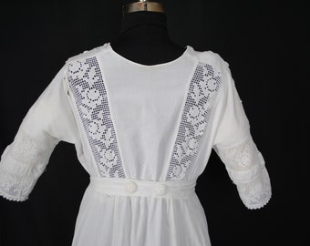 Puede incluir: Vestido blanco vintage con intrincados detalles de encaje en las mangas y la espalda. El vestido presenta una cintura entallada con dos botones blancos y una falda fluida. Las mangas son de largo codo.