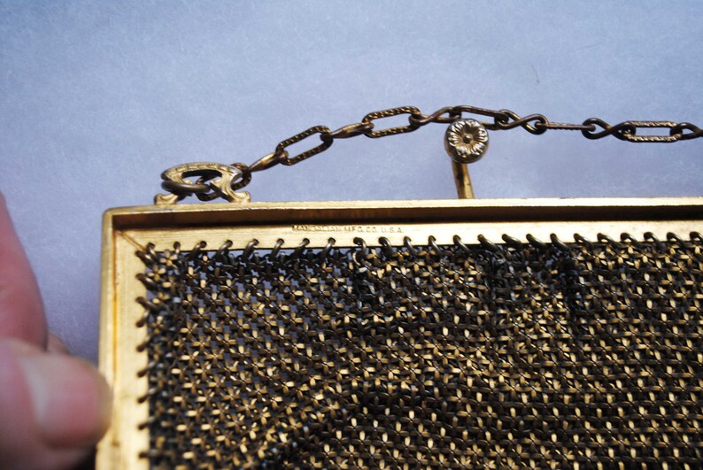 May include: Close-up of a vintage gold-toned mesh purse with a chain handle. The purse has a rectangular frame and a delicate, woven mesh design. The chain and frame show signs of age, adding to its antique charm. The text "MANHATTAN MFG CO. U.S.A." is visible.