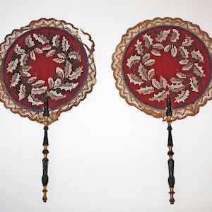 May include: Two antique hand fans. Each fan features a red centre with a wreath of leaves in white, brown, and grey. They have a lace trim and ornate black and gold handles. The fans are decorative.