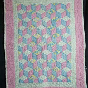 Op de afbeelding: Een handgemaakte babyquilt met een geometrisch patroon van roze, blauwe, witte en gele blokken. De quilt heeft een roze rand en een witte binnenrand. De blokken lijken 3D-kubussen te zijn. De quilt is gemaakt van zachte stof.