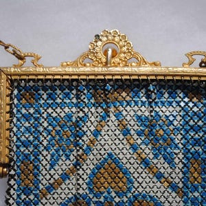 May include: A vintage gold-toned metal mesh purse with a decorative frame and chain. The purse features a geometric pattern in blue, white, and gold. The frame has ornate detailing, and the chain is attached at the top.