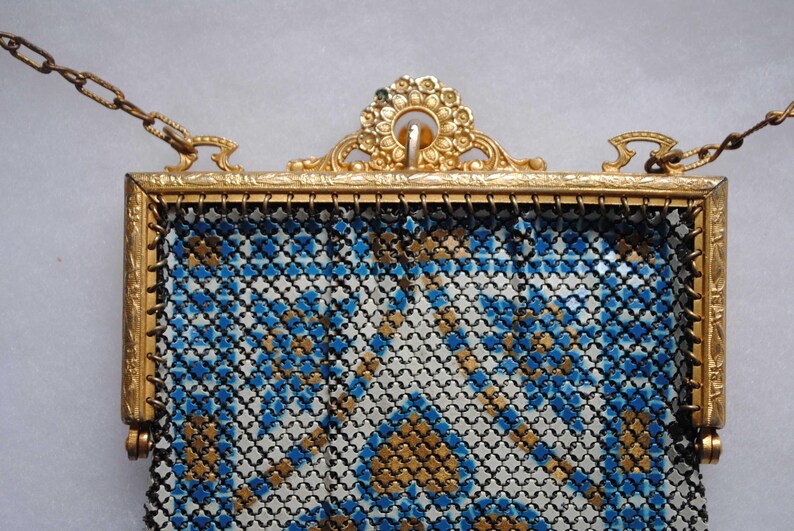 May include: A vintage gold-toned metal mesh purse with a decorative frame and chain. The purse features a geometric pattern in blue, white, and gold. The frame has ornate detailing, and the chain is attached at the top.