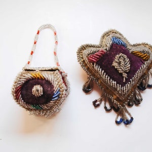 May include: A beaded purse with a handle and a beaded brooch. The purse is round with a dark purple center and colorful accents. The brooch is heart-shaped with a purple center and beaded details.