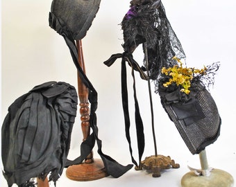 May include: Four antique black hats on display. The hats vary in style, including bonnets and brimmed hats, and are adorned with ribbons, lace, and floral accents. Each hat is mounted on a decorative stand, showcasing their intricate designs and textures.