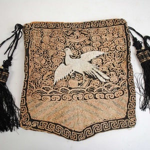 Pode incluir: Uma bolsa retangular ornamentada com um desenho de pássaro. A bolsa apresenta um pássaro branco sobre um fundo preto, rodeado por padrões decorativos bege e preto. Borlas e cordões pretos completam o design.