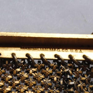 May include: Close-up of a vintage gold-toned metal purse frame. The frame features a floral mesh design and the text "MANDALIAN MFG. CO. U.S.A." is visible. The purse frame has decorative accents and a clasp.