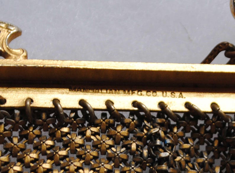 May include: Close-up of a vintage gold-toned metal purse frame. The frame features a floral mesh design and the text "MANDALIAN MFG. CO. U.S.A." is visible. The purse frame has decorative accents and a clasp.