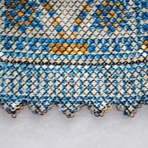 May include: Close-up of a section of a vintage chainmail purse. The purse features a pattern of blue and gold squares on a silver background. The bottom edge has a scalloped design.