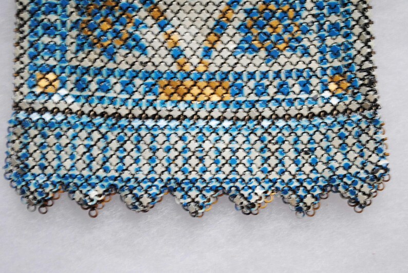 May include: Close-up of a section of a vintage chainmail purse. The purse features a pattern of blue and gold squares on a silver background. The bottom edge has a scalloped design.