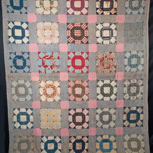 Puede incluir: Un edredón vintage con un diseño de patchwork. El edredón presenta un patrón de cuadrícula de cuadrados, cada uno con un cuadrado central rodeado por un borde contrastante. Los cuadrados son de varios colores y patrones, incluyendo diseños florales y geométricos, con cuadrados rosas entre ellos.