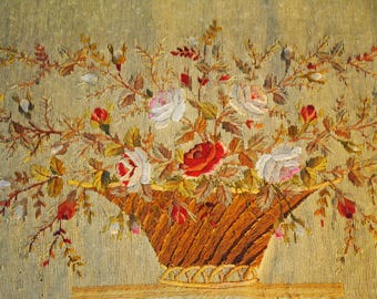 May include: A needlepoint tapestry featuring a brown woven basket filled with a vibrant floral arrangement of red, white, and pink roses, surrounded by green and brown foliage, all set against a textured, light beige fabric background.