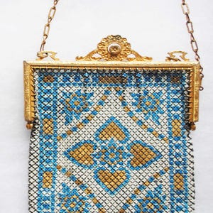 May include: An antique gold-toned metal mesh purse with a chain handle. The purse features a geometric pattern in blue, white, and gold. The frame is ornate with a floral design and a small gemstone.