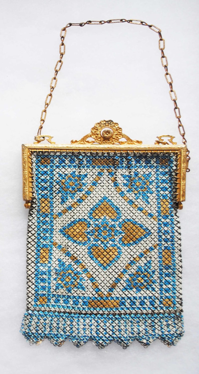 May include: An antique gold-toned metal mesh purse with a chain handle. The purse features a geometric pattern in blue, white, and gold. The frame is ornate with a floral design and a small gemstone.