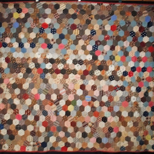 Op de afbeelding: Een vintage quilt met een patchwork-ontwerp. De quilt bestaat uit talrijke zeshoekige stukken stof in verschillende kleuren en patronen, waaronder bruin, blauw, rood en roze. Het heeft een rode rand.