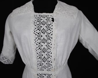 Puede incluir: Vestido blanco vintage con intrincados detalles de encaje. El vestido presenta un escote cuadrado y un panel de encaje decorativo en la parte delantera. Las mangas son de tres cuartos con puños de encaje. Los botones adornan el corpiño y el vestido tiene una cintura entallada.