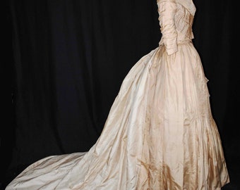 May include: A vintage ivory wedding dress with a long train, displayed on a mannequin. The dress features a fitted bodice with detailed sleeves and a full skirt. The fabric has a subtle sheen, and the train extends several feet.
