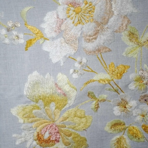Könnte beinhalten: Nahaufnahme eines gestickten Blumendesigns. Das Design zeigt große weiße und gelbe Blumen mit rosa und braunen Akzenten, zusammen mit kleineren weißen Blumen und gelben Blättern, alles auf einem hellblauen Stoffhintergrund.