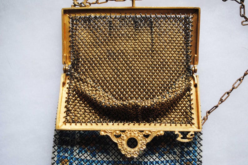 May include: An open, antique gold-toned metal mesh purse with a decorative frame and chain handle. The purse features a detailed, ornate clasp and a blue and white patterned lining visible at the bottom.