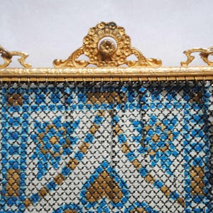May include: Vintage gold-toned metal mesh purse with a decorative frame and chain handle. The purse features a geometric pattern in blue, gold, and white. The frame has ornate detailing, adding to its antique charm. A classic accessory.