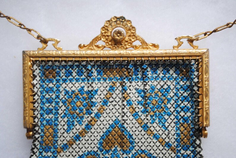 May include: Vintage gold-toned metal mesh purse with a decorative frame and chain handle. The purse features a geometric pattern in blue, gold, and white. The frame has ornate detailing, adding to its antique charm. A classic accessory.