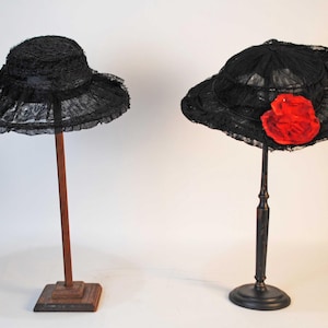 Peut inclure: Deux chapeaux en dentelle noire sur des présentoirs. L'un a un large bord et un design simple. L'autre chapeau présente une grande fleur rouge sur le côté. Les chapeaux sont exposés sur des présentoirs en bois et en métal.