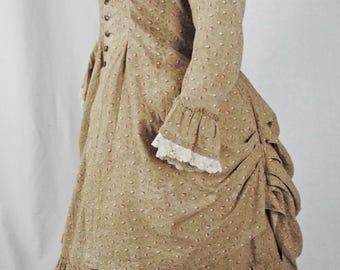 May include: A tan-coloured, vintage-style dress with a small pink floral pattern. The dress features long sleeves with lace cuffs, a high collar, and a tiered, ruffled skirt. The dress is buttoned down the front.