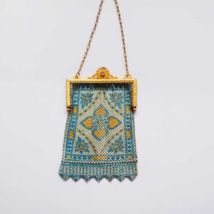 May include: An antique gold-toned metal mesh purse with a decorative frame and chain handle. The purse features a blue and yellow geometric pattern, with a scalloped edge at the bottom. The purse is suspended against a white background.
