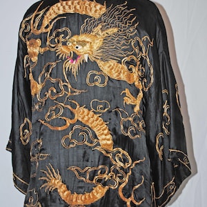 Pode incluir: Robe preta com bordado de dragão dourado. O design do dragão apresenta detalhes intrincados, incluindo escamas e nuvens. A robe é exibida em um manequim, mostrando a forma e o design da peça.