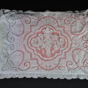 Puede incluir: Una funda de almohada rectangular blanca con intrincados bordados calados y un motivo floral central. La tela está adornada con elaborados patrones de volutas y terminada con un delicado borde de encaje festoneado en los cuatro lados.