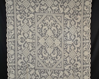 May include: Cream-colored lace tablecloth with an intricate floral and geometric design. The rectangular cloth features a central panel with diamond and leaf patterns, surrounded by a detailed border. The edges have a scalloped trim.