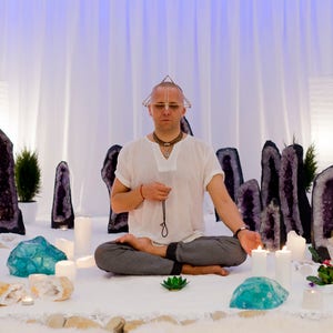 May include: A man sits cross-legged on a white surface surrounded by amethyst geodes and candles. He is wearing a white shirt and a crystal pyramid sits on his head.