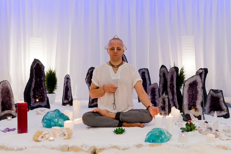 May include: A man sits cross-legged on a white surface surrounded by amethyst geodes and candles. He is wearing a white shirt and a crystal pyramid sits on his head.