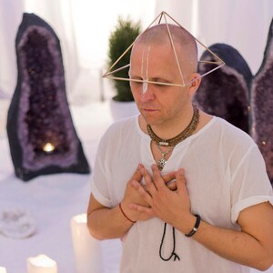 May include: A man wearing a white shirt and a gold pyramid-shaped headpiece is meditating with his hands clasped over his heart. He is surrounded by large amethyst crystals and candles.