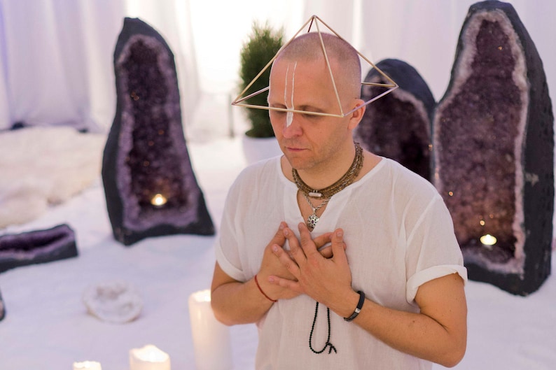 May include: A man wearing a white shirt and a gold pyramid-shaped headpiece is meditating with his hands clasped over his heart. He is surrounded by large amethyst crystals and candles.