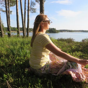 May include: A woman wearing sunglasses sits in a field of green grass with a lake in the background. She is wearing a yellow and white striped shirt and a pink and white patterned skirt. She has a geometric wire headpiece on her head.