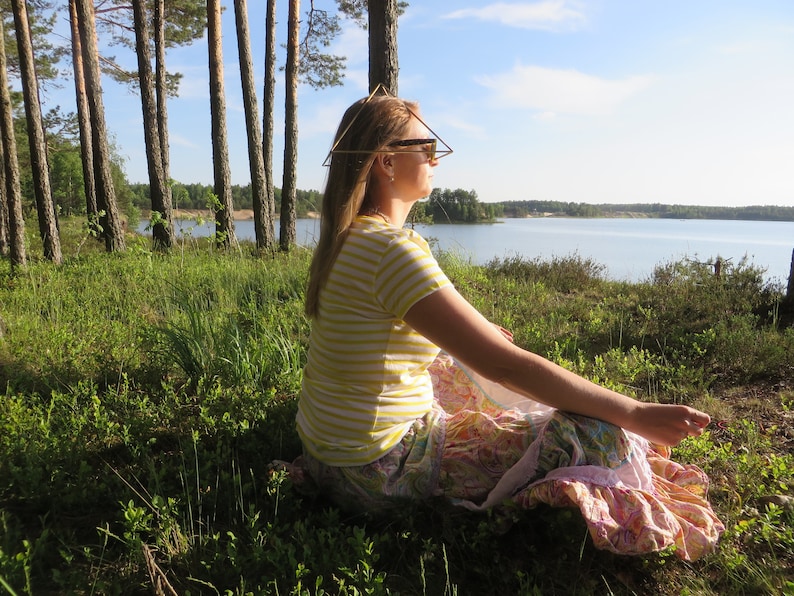 May include: A woman wearing sunglasses sits in a field of green grass with a lake in the background. She is wearing a yellow and white striped shirt and a pink and white patterned skirt. She has a geometric wire headpiece on her head.