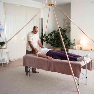 May include: A large, hollow copper pyramid frame constructed from thin, straight metal tubing. The structure features four long poles meeting at a central apex point, designed to stand over a massage table for use in a room setting.