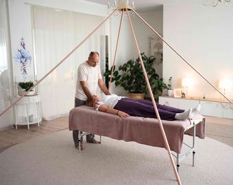 May include: A large, hollow copper pyramid frame constructed from thin, straight metal tubing. The structure features four long poles meeting at a central apex point, designed to stand over a massage table for use in a room setting.