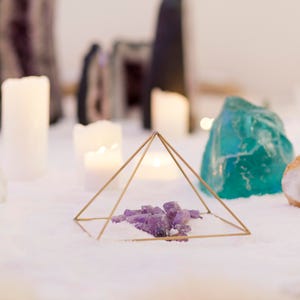 May include: A gold geometric pyramid sits on a white surface with a cluster of purple amethyst crystals inside. The pyramid is surrounded by white and teal crystals, candles, and a white surface.