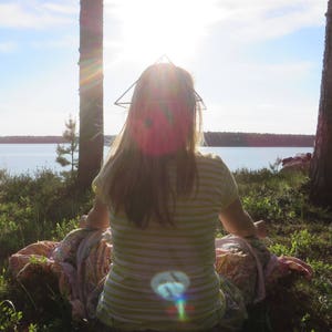 May include: A woman sits on a blanket in a forest clearing, wearing a striped shirt and a geometric wire headpiece. The sun shines brightly behind her, creating a lens flare effect.