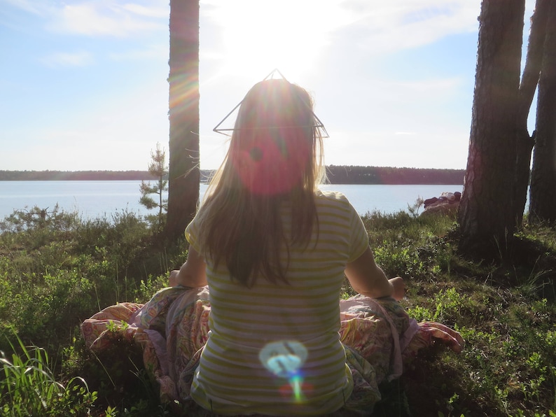 May include: A woman sits on a blanket in a forest clearing, wearing a striped shirt and a geometric wire headpiece. The sun shines brightly behind her, creating a lens flare effect.