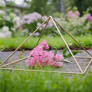 May include: A gold geometric pyramid frame sits on a bed of green grass with a bouquet of pink flowers inside.