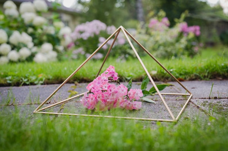 May include: A gold geometric pyramid frame sits on a bed of green grass with a bouquet of pink flowers inside.