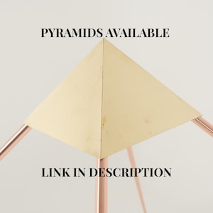 Könnte beinhalten: Eine geometrische Skulptur mit einer goldenen Pyramidenoberseite und kupferfarbenen Beinen. Der Text "PYRAMIDS AVAILABLE" und "LINK IN DESCRIPTION" wird in schwarzer Schrift angezeigt. Die Pyramide ist zentriert, wobei sich die Beine nach außen erstrecken.