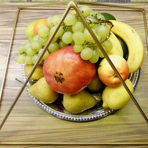 May include: A gold metal pyramid frame sits on a wooden surface, holding a silver bowl filled with fruit. The fruit includes green grapes, a red pomegranate, yellow pears, a yellow lemon, a red apple, and a yellow banana.