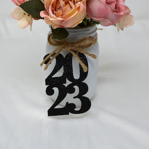 May include: A white mason jar with a burlap bow and a black glitter 2023 sign. The jar is filled with a bouquet of pink and peach artificial flowers.