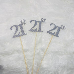 May include: Three silver glitter cake toppers with the number 21 and the word "st" on each. The toppers are on wooden sticks.