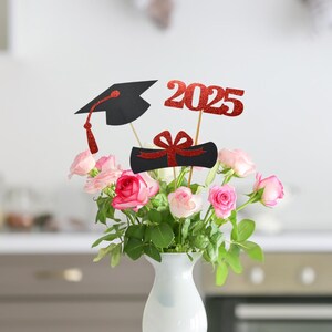 Puede incluir: Centro de mesa para una fiesta de graduaci&oacute;n con rosas rosadas en un jarr&oacute;n blanco. El centro de mesa incluye una gorra de graduaci&oacute;n negra, un diploma rojo con un lazo y un 2025 de purpurina roja.